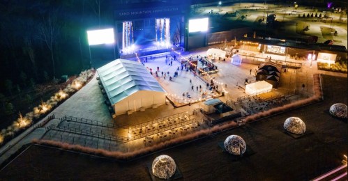 Aerial view of an outdoor event with a stage, tents, and seating, surrounded by illuminated pathways and domes.