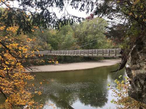 A wooden suspension bridge spans a calm river, surrounded by autumn foliage and rocky cliffs.