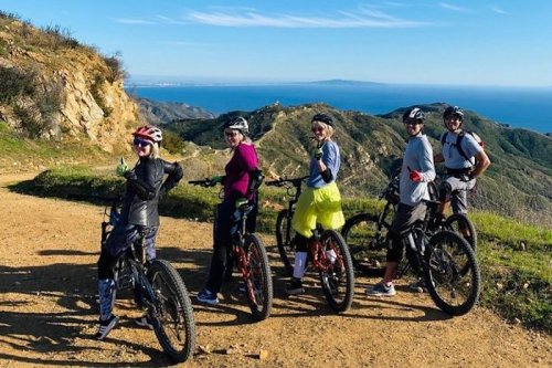 A group of five cyclists poses on a scenic mountain trail with ocean views in the background.