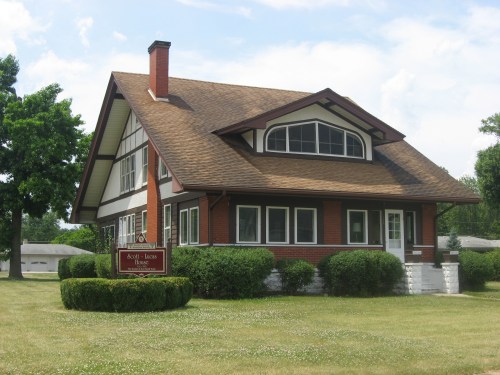 A large, two-story house with a brown roof, red brick, and white trim, surrounded by green grass and bushes.