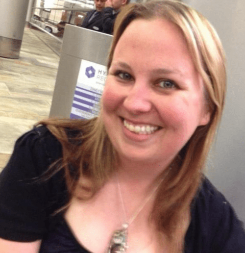 A smiling woman with long, light brown hair, wearing a black top, poses for a photo in a public space.