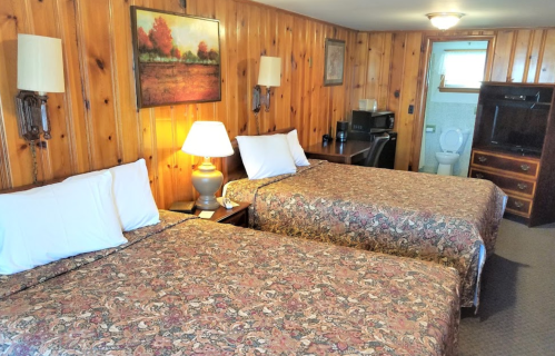 A cozy room with two beds, wooden walls, a lamp, and a desk, featuring a bathroom in the background.