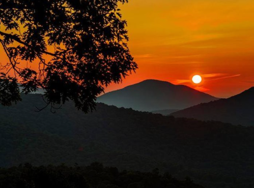 A vibrant sunset over rolling mountains, with silhouettes of trees in the foreground and warm orange hues in the sky.