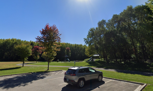 A sunny park scene with a gray car parked in the foreground and trees with autumn colors in the background.