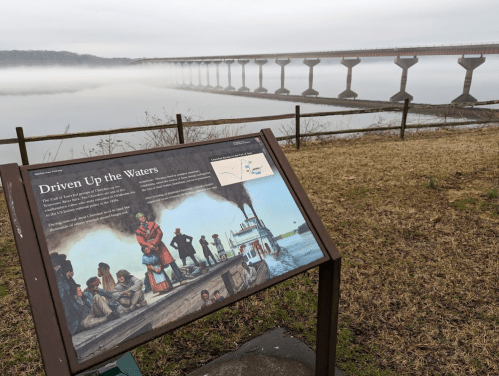 A historical sign titled "Driven Up the Waters" near a foggy river with a bridge in the background.