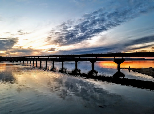 A bridge spans a calm river at sunset, reflecting vibrant colors in the water beneath a cloudy sky.