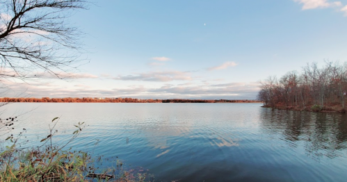 A serene lake scene with calm water, trees lining the shore, and a clear blue sky with scattered clouds.