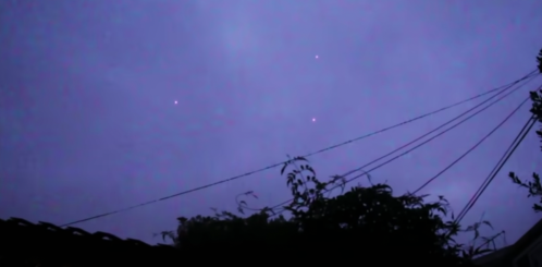 Three bright points of light appear in a purple twilight sky, with silhouettes of trees and power lines in the foreground.