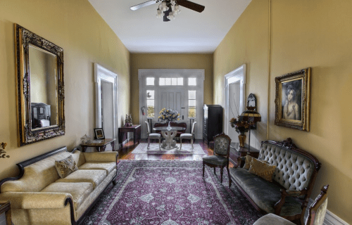 A spacious, elegantly decorated living room with vintage furniture, mirrors, and a patterned rug.