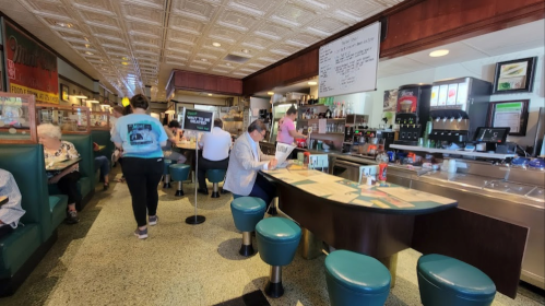 A bustling diner interior with teal seating, patrons at tables, and a counter with staff serving food and drinks.