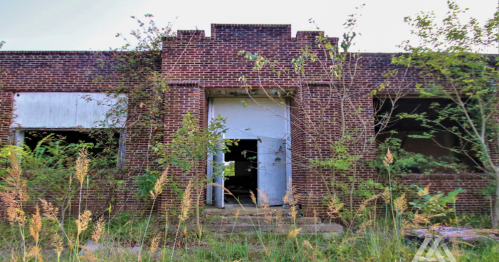 Abandoned brick building with overgrown grass and plants, featuring boarded windows and a central doorway.
