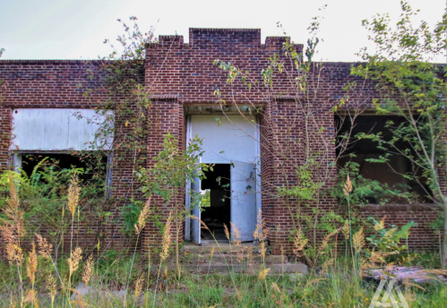 Abandoned brick building with overgrown grass and plants, featuring boarded windows and a central doorway.