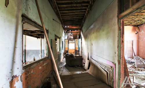 Abandoned hallway with peeling paint, exposed beams, and debris scattered on the floor. A sofa is visible in the distance.