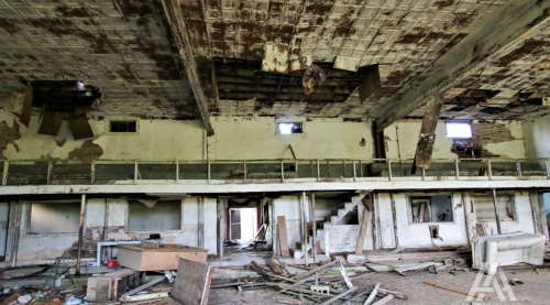 Interior of an abandoned building with a damaged ceiling, debris on the floor, and broken windows.