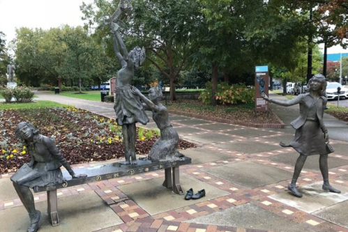 A bronze sculpture depicting three girls in playful poses, set in a park with trees and flowers in the background.