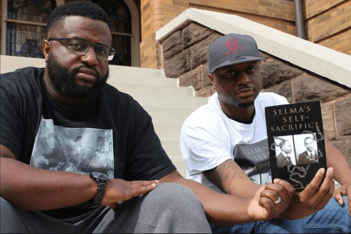 Two men sitting on steps, one holding a book titled "Selma's Self-Sacrifice," both looking at the camera.