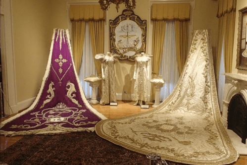 Two ornate capes displayed in a room, one purple with gold embroidery and the other cream with intricate designs.
