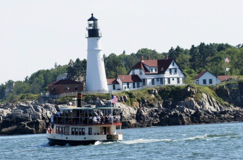 A boat sails past a white lighthouse and a coastal house on a sunny day.