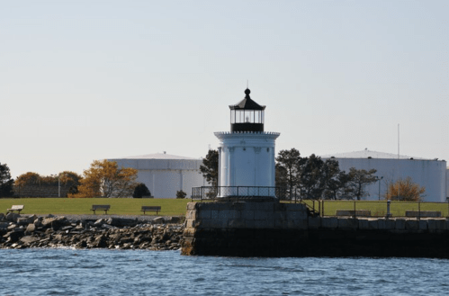 A white lighthouse stands on a rocky shore, with green grass and storage tanks in the background under a clear sky.