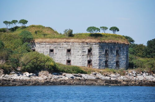 Abandoned stone structure on a grassy island, surrounded by water and rocky shoreline under a clear blue sky.