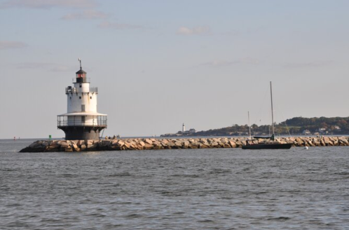 A lighthouse stands on a rocky pier, with a sailboat nearby and a distant shoreline under a clear sky.