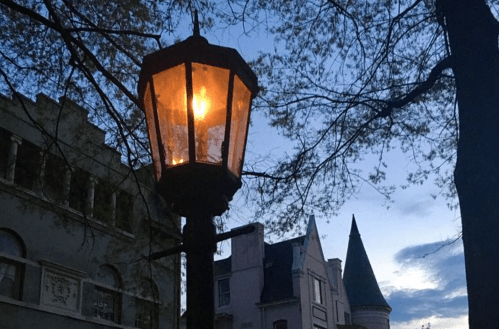 A glowing street lamp stands against a twilight sky, with trees and historic buildings in the background.