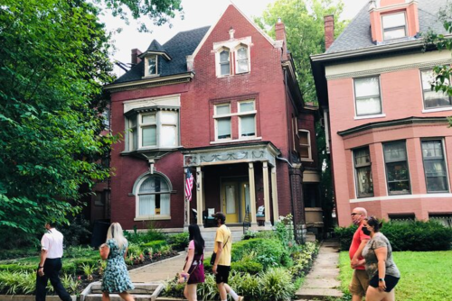 A red brick house with a porch, surrounded by greenery, as people walk by on a sunny day.