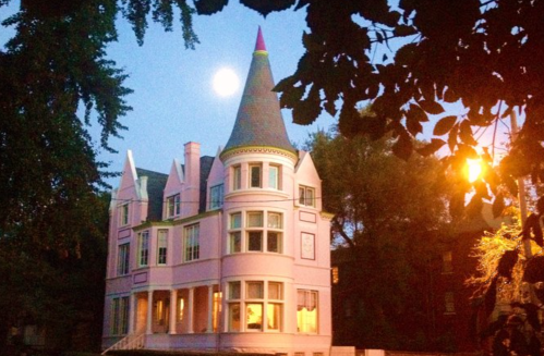 A pink Victorian house with a pointed tower, illuminated by moonlight, surrounded by trees at dusk.