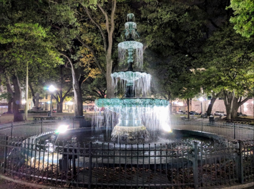 A illuminated three-tiered fountain at night, surrounded by trees and a black fence in a quiet park.