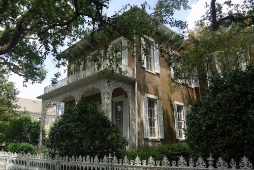 A historic two-story house with intricate white trim, surrounded by lush greenery and a white picket fence.