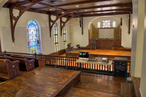 Interior of a spacious hall with wooden pews, stained glass windows, and an organ at the front. Equipment is set up for recording.