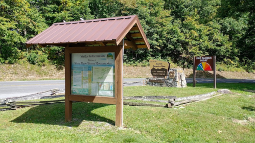 Information sign at a visitor center, featuring maps and directions, surrounded by greenery and a road.