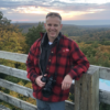 A man in a red plaid jacket stands by a wooden railing, holding a camera, with a scenic landscape in the background.