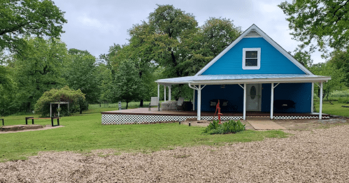 A blue house with a porch surrounded by green trees and a gravel path in a serene outdoor setting.