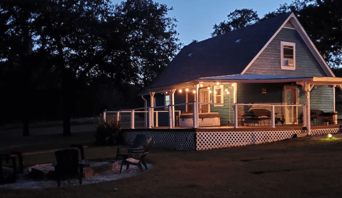 A cozy blue house at dusk, with a wraparound porch and a fire pit surrounded by chairs in the foreground.