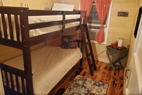 A cozy bedroom featuring a wooden bunk bed, a colorful pillow, a small table, and a rug on a wooden floor.