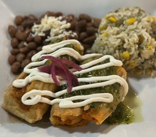 A plate featuring two tamales topped with green sauce and sour cream, served with rice and beans.