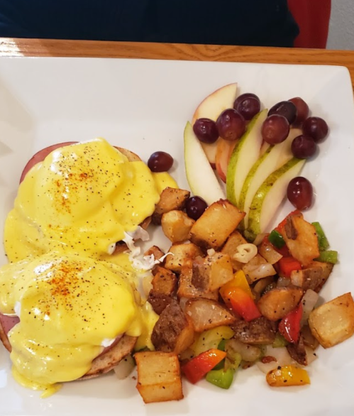 A plate with eggs Benedict, seasoned potatoes, and a side of sliced apples and grapes.