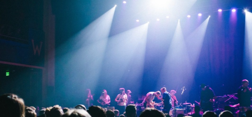 A lively concert scene with a band performing on stage, illuminated by bright lights, and an engaged audience in front.