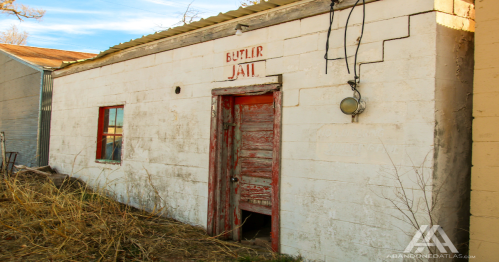 Abandoned building with a weathered sign reading "Butler Jail," featuring a red door and overgrown grass.