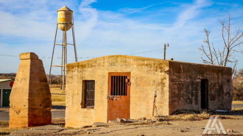 An old, weathered jail building with barred windows, next to a tall water tower against a clear blue sky.