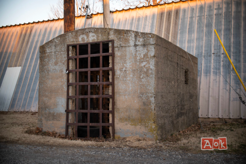 A weathered concrete structure with a barred door, set against a backdrop of a metal building and gravel ground.