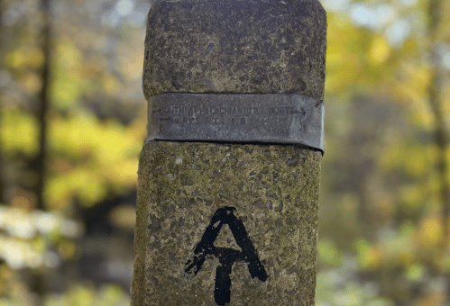 A weathered stone marker with an engraved plaque and a black letter "A" painted on it, surrounded by autumn foliage.
