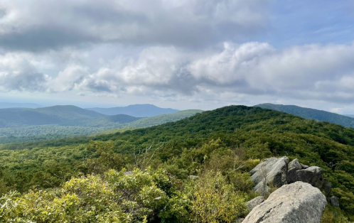 A panoramic view of lush green mountains under a cloudy sky, with rocky outcrops in the foreground.