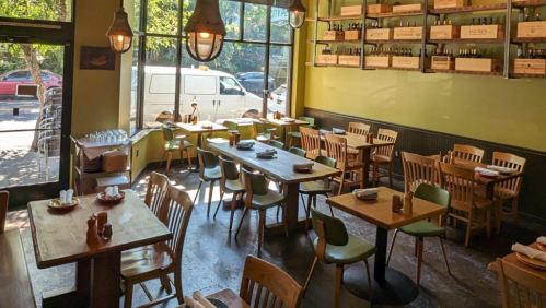 Cozy restaurant interior with wooden tables, chairs, and large windows letting in natural light.