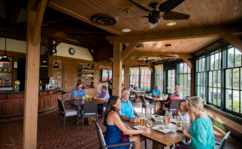 A cozy restaurant interior with wooden beams, large windows, and diners enjoying meals and drinks at tables.