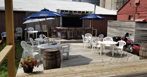Outdoor seating area with white chairs, tables, umbrellas, and a motorcycle, surrounded by wooden structures and greenery.