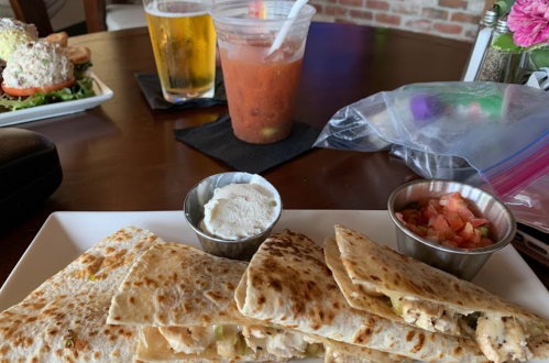 A plate of quesadillas with dipping sauces, a cocktail, and a salad in the background on a wooden table.
