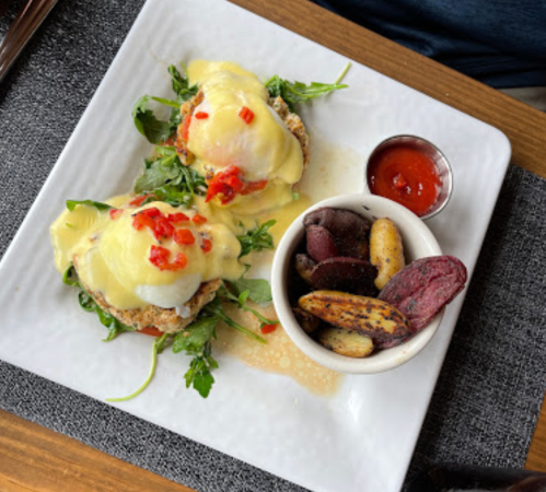 Two eggs Benedict on a bed of greens, topped with hollandaise sauce and diced peppers, served with a side of roasted potatoes.