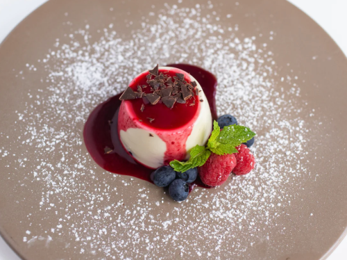 A creamy dessert topped with berry sauce, chocolate shavings, and fresh raspberries and blueberries on a plate.